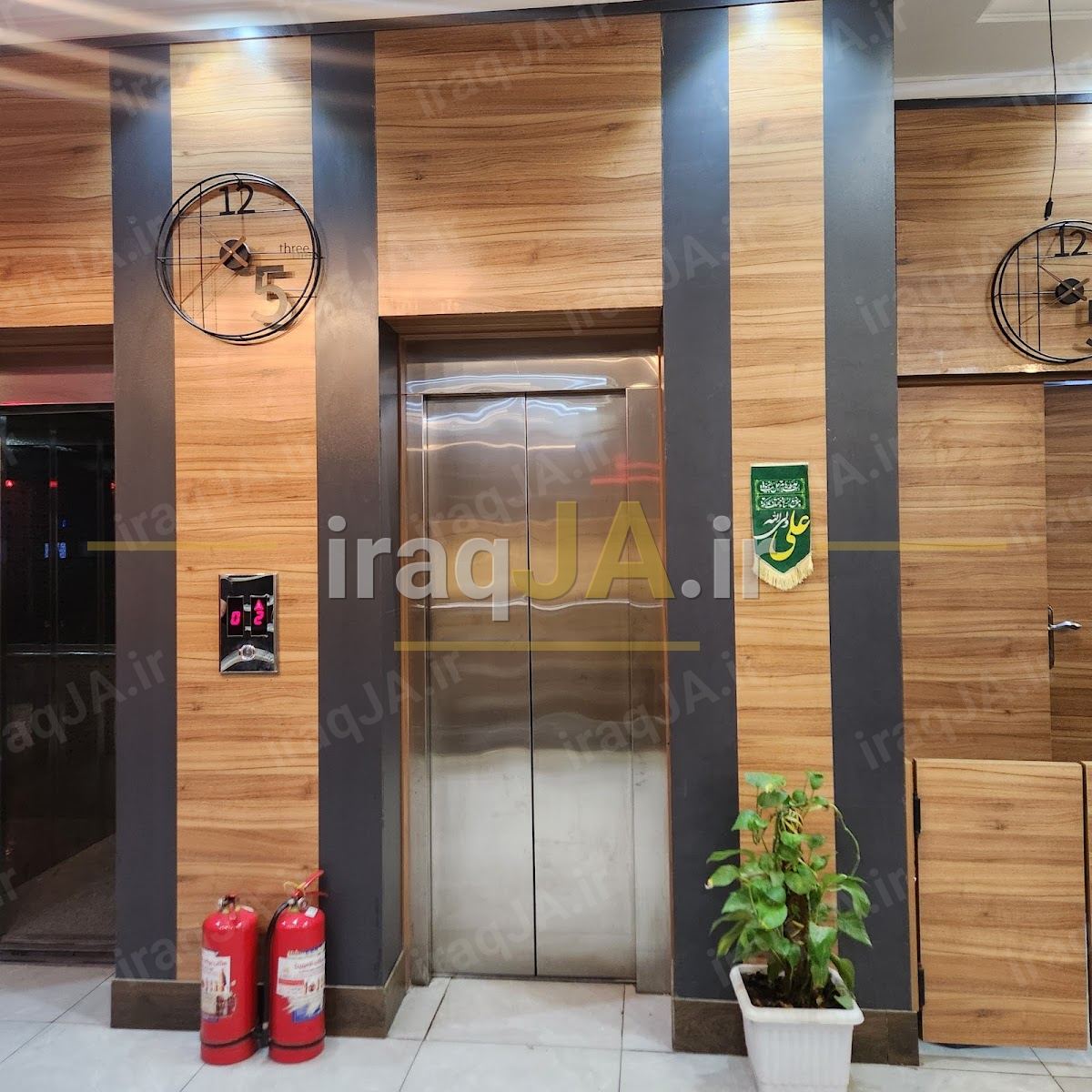 محوطهٔ آسانسور با دیوارکوبِ چوبی، ساعتِ دیواری و کپسولِ آتش‌نشانی