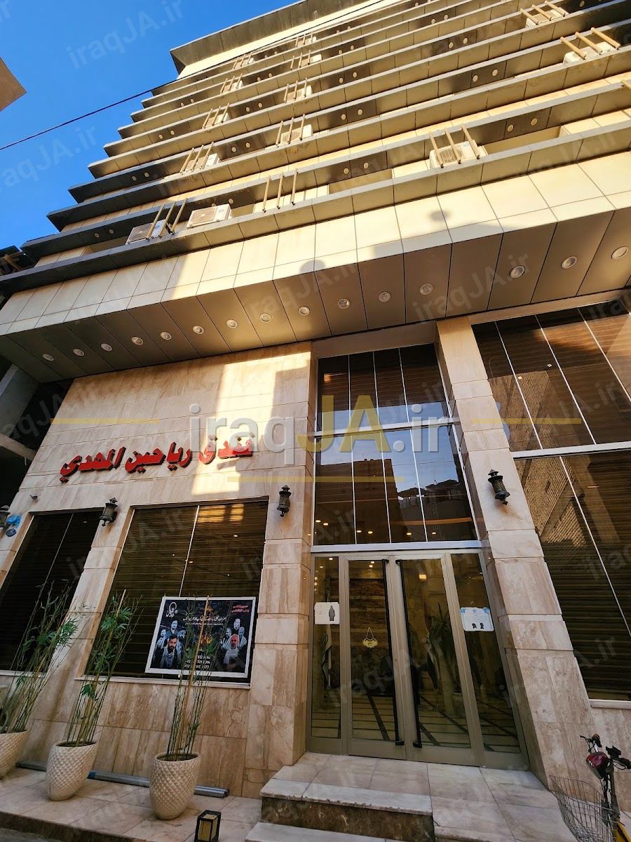 ورودیِ هتل با تابلوی عربیِ «فندق رياحين المهدي» و گلدان‌های دو سویِ در