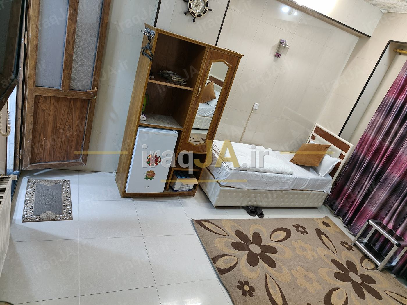 اتاق ساده با تختِ یک‌نفره، کمدِ چوبیِ یخچال‌دار و قالیچهٔ گل‌دار